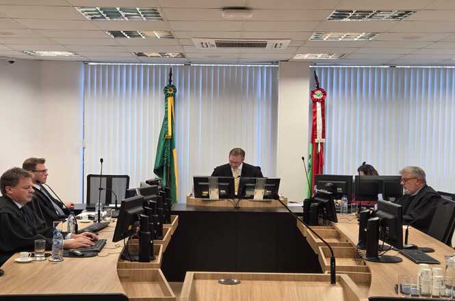 A imagem mostra uma sala de tribunal com cinco pessoas sentadas em bancadas. No centro, há uma pessoa em destaque, provavelmente o juiz. As outras pessoas estão sentadas ao redor em bancadas equipadas com computadores e microfones. No fundo, há duas bandeiras: a do Brasil e outra que parece ser de um estado ou instituição. A sala tem janelas grandes com persianas verticais fechadas, e o ambiente é formal e organizado.