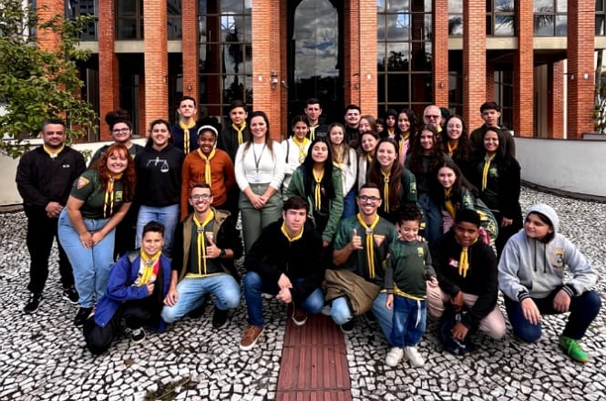A imagem mostra um grupo de pessoas posando em frente a um edifício de tijolos vermelhos com grandes janelas de vidro. As pessoas estão em um caminho pavimentado com pedras decorativas e muitas delas estão usando uniformes com blusas verdes e lenços amarelos, sugerindo que podem fazer parte de um grupo ou organização específica. O fundo revela uma arquitetura moderna e o clima parece estar ameno ou levemente nublado. As expressões faciais transmitem  descontração e alegria.