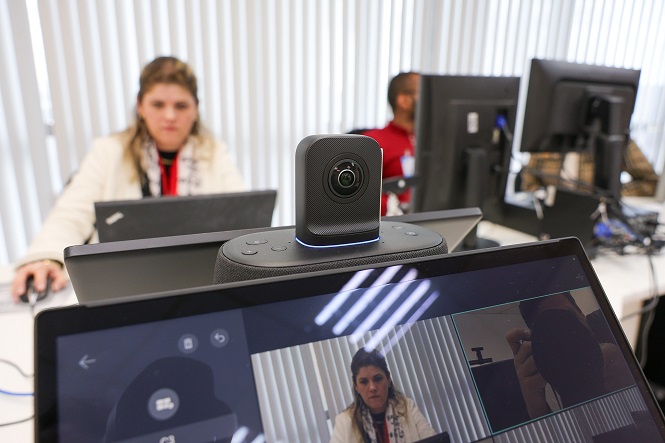A imagem mostra um ambiente de escritório com pessoas trabalhando em computadores. No centro, há uma câmera de videoconferência ligada a um dispositivo posicionado sobre um monitor. No monitor, a tela exibe a imagem da pessoa à frente, capturada pela câmera. Atrás, é possível ver duas pessoas focadas em suas telas de computador. As janelas no fundo deixam entrar luz natural, iluminando o espaço.