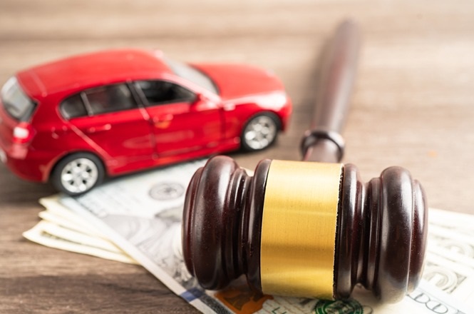 A imagem mostra um martelo de juiz, algumas notas de dólar e um carro em miniatura vermelho. O martelo está sobre as notas, sugerindo uma representação de questões legais ou jurídicas relacionadas a finanças ou automóveis. A imagem transmite uma associação entre a justiça, dinheiro e automóveis.