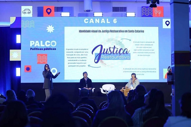 A imagem mostra um evento em um palco de conferência ou seminário. Há uma grande tela ao fundo com o texto "Justiça Restaurativa" e informações adicionais sobre o tema, incluindo "Identidade visual da Justiça Restaurativa em Santa Catarina". Três pessoas estão no palco: uma pessoa em pé, provavelmente fazendo uma apresentação, e duas pessoas sentadas, possivelmente painelistas ou palestrantes. No canto esquerdo da tela, lê-se "PALCO Políticas públicas". O público está sentado em cadeiras no espaço do evento.