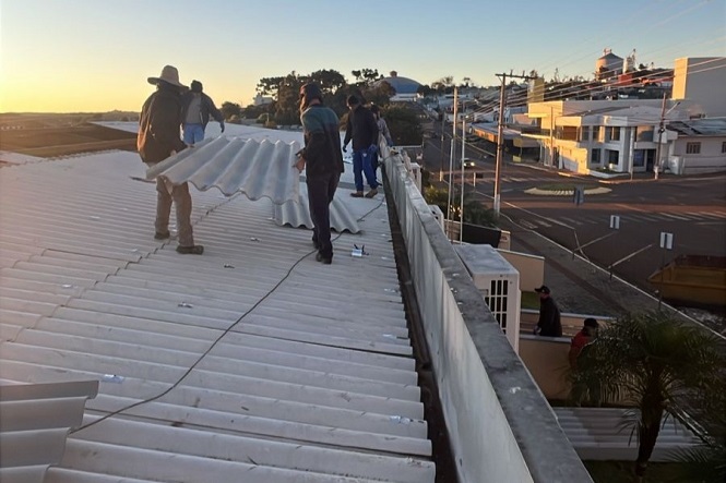 A imagem mostra um grupo de pessoas trabalhando em um telhado. Elas parecem estar instalando ou reparando telhas de material claro. O telhado é plano e há visão de uma rua e prédios ao fundo, sob luz do fim de tarde ou amanhecer. Algumas pessoas estão carregando uma telha grande, enquanto outras as acompanham. O ambiente parece ser uma área urbana.