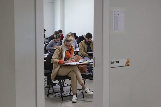 A imagem mostra uma sala de aula onde várias pessoas estão sentadas em carteiras, aparentemente realizando uma prova ou concentradas em atividades escritas. Ao centro, uma pessoa loira está vestindo um casaco bege e um cachecol laranja, escrevendo em papéis sobre a mesa. Outras pessoas ao fundo parecem estar igualmente focadas em suas tarefas. A imagem é tirada de fora da sala e a porta está aberta, revelando a cena interna. Na porta, há uma placa indicando o número da sala, "C 104".