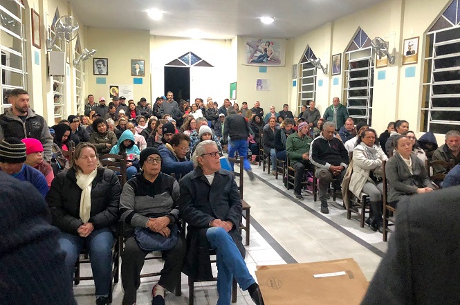 A imagem mostra uma sala cheia de pessoas sentadas em cadeiras, aparentemente participando de uma reunião ou evento comunitário. Há homens e mulheres de várias idades, todos voltados para a frente da sala. As paredes estão decoradas com quadros e há janelas grandes que permitem a entrada de luz. O ambiente parece ser informal e acolhedor.