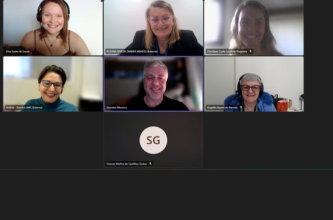 A imagem mostra uma captura de tela de uma videoconferência com sete participantes. Seis pessoas estão visíveis, sorrindo e olhando para a câmera, enquanto uma delas é representada apenas por iniciais ("SG") em um fundo preto. As janelas dos participantes estão organizadas em uma grade, comum em plataformas de reunião online. Cada janela também exibe nomes, sugerindo que os participantes estão identificados. O pano de fundo de cada participante parece ser um ambiente interno, com boa iluminação.