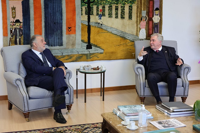 A imagem mostra dois homens sentados em poltronas cinzas em uma sala. Elas estão conversando e parecem estar em um ambiente formal. Atrás delas, há uma tapeçaria colorida pendurada na parede, que retrata uma cena de uma rua. No chão, há um tapete em tons de vermelho e azul. Há uma mesa de centro com livros e xícaras de café à frente das duas pessoas. O ambiente é bem iluminado e organizado.