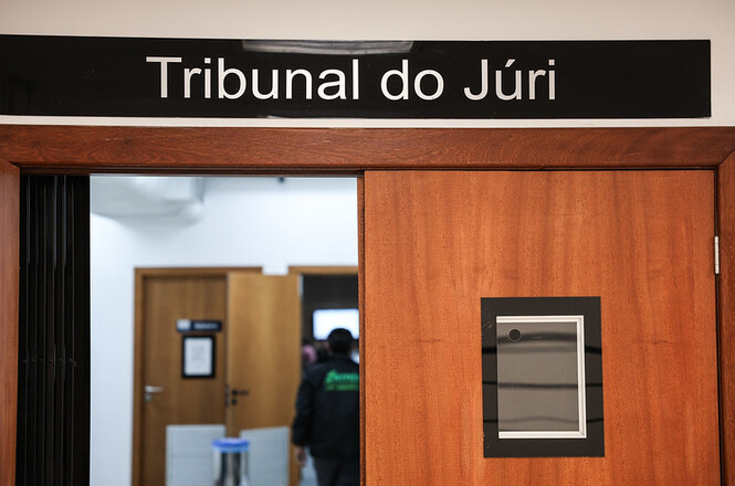 A imagem mostra uma porta com uma placa preta acima dela que diz "Tribunal do Júri". A porta é de madeira e tem um retângulo preto com um quadrado cinza no interior. No fundo, há outra porta e algumas pessoas entrando ou saindo do espaço. O ambiente parece ser parte de um edifício judicial.