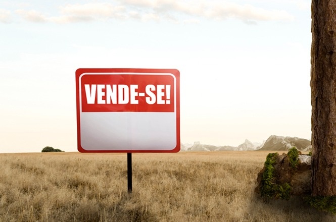 A imagem mostra uma placa vermelha com bordas brancas no centro de um campo gramado, com as palavras “VENDE-SE!” em destaque. Ao fundo, há uma paisagem ampla com montanhas ao longe e um céu claro. À direita da imagem, é visível parte de um tronco de árvore.