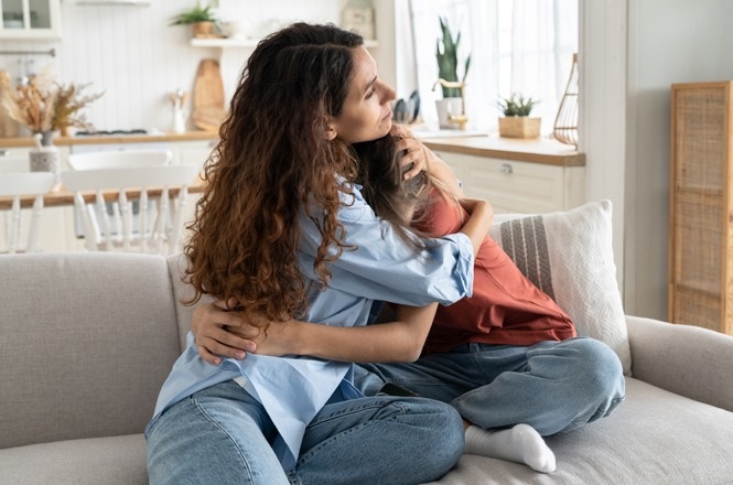 A imagem mostra duas pessoas sentadas em um sofá, abraçando-se de forma afetuosa. Uma pessoa está usando uma camisa azul clara e calças jeans, enquanto a outra está vestindo uma camiseta marrom-avermelhada e também calças jeans. O ambiente ao fundo parece ser uma sala de estar com decoração acolhedora, incluindo cadeiras e elementos decorativos.