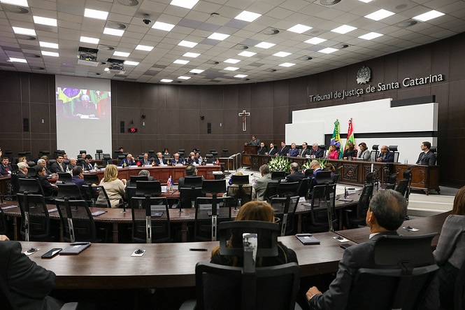 A imagem mostra uma sala de audiências no "Tribunal de Justiça de Santa Catarina". Há várias pessoas sentadas ao redor de uma grande mesa em formato de "U". No centro da sala, há um púlpito com algumas pessoas se dirigindo ao público. Há bandeiras do Brasil e de Santa Catarina, além de um crucifixo na parede. O ambiente tem um telhado com várias luzes embutidas e um sistema de projeção ao fundo. Ao lado direito, um grupo maior de pessoas está posicionado em um espaço elevado.