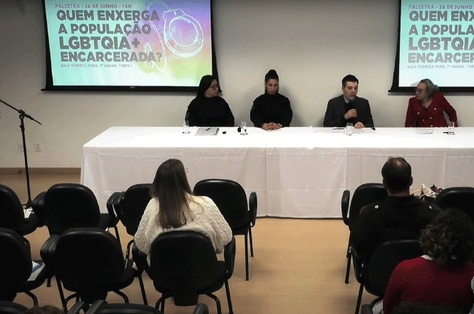 A imagem mostra uma mesa de debate com quatro pessoas sentadas à frente de um público em um auditório. O evento trata de temas relacionados à população LGBTQIA+ encarcerada, conforme indicado no telão ao fundo, que diz: "Quem enxerga a população LGBTQIA+ encarcerada?". A plateia está sentada em cadeiras, assistindo à palestra ou discussão que está ocorrendo. Há um microfone posicionado à esquerda da imagem, próximo à mesa onde os palestrantes estão sentados.