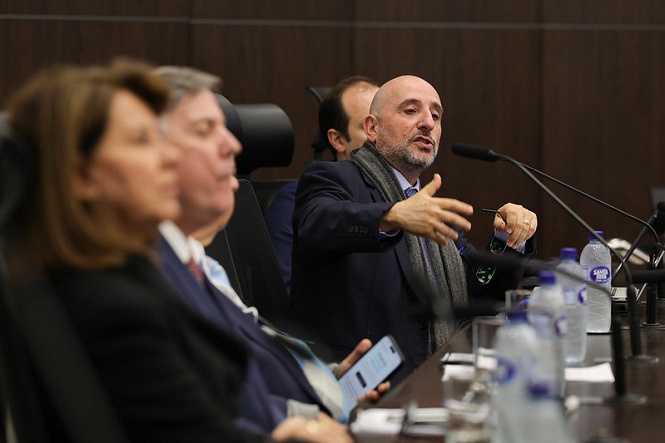 Na imagem, há quatro pessoas sentadas em uma mesa, participando de uma reunião ou conferência. Todos estão vestidos formalmente. Um dos homens está gesticulando com a mão, enfatizando sua fala. Na mesa, há microfones e várias garrafas de água. A cena parece estar em um ambiente formal, possivelmente uma sala de conferências ou um auditório.