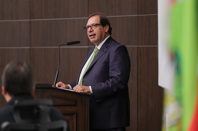 Na imagem, há um homem de terno e gravata verde, falando em pé atrás de um púlpito com um microfone. Ele parece estar em um ambiente formal, possivelmente durante um discurso ou apresentação. No fundo, há uma parede de madeira e parte de uma bandeira. Outras pessoas estão presentes, mas estão desfocadas na imagem.
