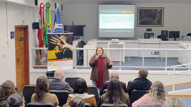 A imagem mostra uma mulher falando em frente a um público em um ambiente que parece ser uma sala de conferências ou auditório. Ela está segurando um microfone. Atrás dela, há uma projeção na tela e bandeiras do Brasil e de um estado ou município. À esquerda, um banner está visível. O público está sentado, prestando atenção à apresentação.