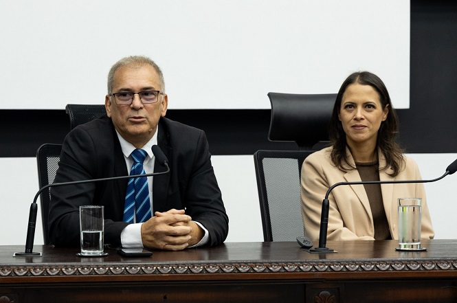 A imagem mostra duas pessoas sentadas em uma mesa de conferência. O homem à esquerda veste um terno escuro e uma gravata azul, e há um copo de água à sua frente. A mulher à direita veste um blazer bege e há também um copo de água à sua frente. Ambos estão em cadeiras de escritório, e há microfones na mesa. O fundo é simples, com uma parede clara e parte de uma tela de projeção visível.
