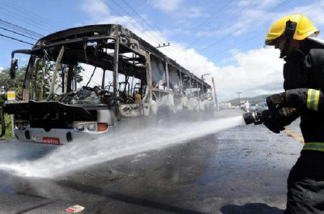 A imagem mostra um ônibus queimado, com danos aparentes em toda a sua estrutura. Ao lado, um bombeiro está usando uma mangueira para apagar o fogo ou resfriar os destroços. O cenário parece ser uma rua durante o dia, com céu parcialmente nublado.