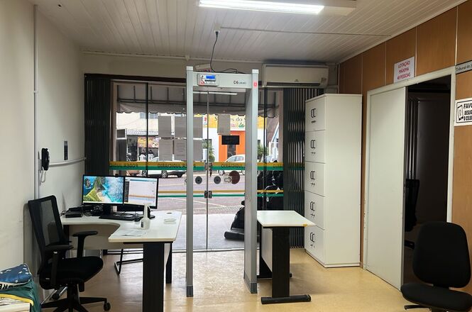 A imagem mostra um interior de um escritório ou recepção. Há uma mesa com um computador e cadeira giratória ao lado esquerdo. No centro, um detector de metais está posicionado na entrada. À direita, há um armário branco com gavetas e uma porta aberta. As paredes são revestidas em madeira clara, e há placas de sinalização. A iluminação é feita com luzes fluorescentes no teto.