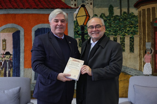 A imagem mostra dois homens sorrindo enquanto seguram um livro juntos. Ambos estão vestidos formalmente. Ao fundo, há uma tapeçaria colorida com cenas de uma paisagem urbana, incluindo casas e árvores, com personagens desenhados. Eles parecem estar em um ambiente fechado, possivelmente um escritório ou sala de reunião.