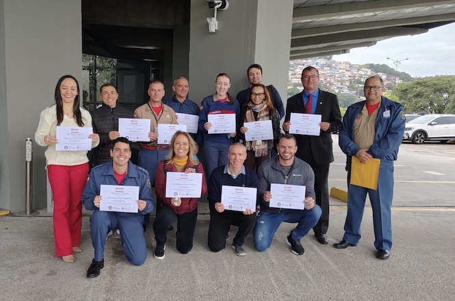 A imagem mostra um grupo de pessoas posando para uma foto. Eles estão segurando certificados, sugerindo que participaram de algum evento ou treinamento. Todos parecem estar em um ambiente externo, possivelmente em frente a um edifício ou estrutura. As pessoas estão sorrindo e parecem orgulhosas de suas conquistas. Há uma variedade de estilos de roupas, desde uniformes até roupas sociais. No fundo, pode-se ver parte de uma cidade ou área urbana.