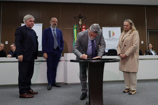 A imagem mostra um grupo de pessoas em um evento formal, provavelmente em ambiente governamental ou judicial. Um homem está assinando um documento em uma mesa no centro. Outros três adultos observam a situação. Ao fundo, há bandeiras e o símbolo do "Poder Judiciário de Santa Catarina". A decoração do ambiente é formal, com paredes de madeira e uma imagem de um crucifixo pendurado, sugerindo ser uma sala de reuniões ou conferências.