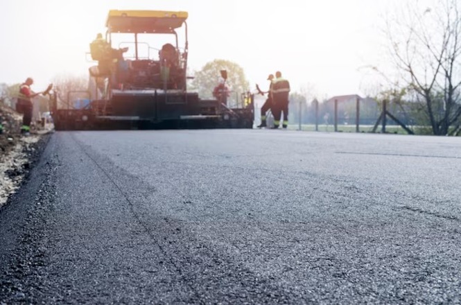 Na imagem, há uma estrada sendo pavimentada. É possível ver uma máquina de pavimentação asfáltica trabalhando, cercada por alguns trabalhadores. O asfalto novo está sendo colocado na superfície da estrada. Ao fundo, há árvores e uma cerca, sugerindo um ambiente ao ar livre. A iluminação indica que pode ser durante o dia.