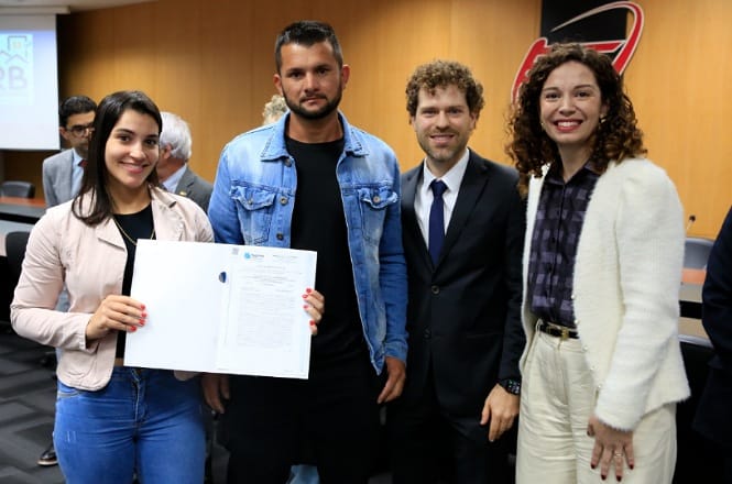 A imagem mostra quatro pessoas posando para uma foto em um ambiente interno que parece ser uma sala de conferências. Uma das pessoas, à esquerda, está segurando um documento aberto, parecendo exibi-lo para a câmera. As outras três pessoas estão ao seu lado, duas delas vestidas de modo formal com ternos, e uma com uma jaqueta jeans. Todos estão sorrindo e aparentam estar participando de um evento oficial. Ao fundo, é possível ver cadeiras e uma parede de madeira.