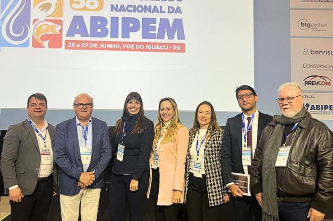 Na imagem, há um grupo de pessoas posando para uma foto em frente a um painel que anuncia o "58º Congresso Nacional da ABIPEM", realizado de 25 a 27 de junho em Foz do Iguaçu, PR. Todos estão vestindo roupas formais e usam crachás de identificação de evento. O ambiente parece ser um auditório ou uma sala de conferência, sugerindo que estão participando de um evento oficial ou profissional.