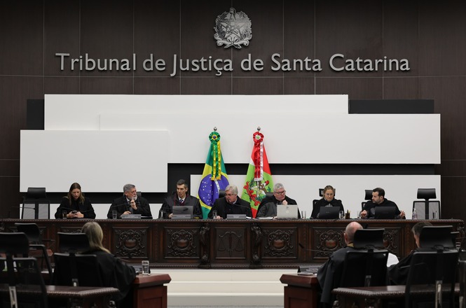 A imagem mostra uma sessão no Tribunal de Justiça de Santa Catarina. Várias pessoas estão sentadas à mesa, provavelmente juízes ou autoridades judiciais, vestidas com togas. Ao fundo, há as bandeiras do Brasil e do estado de Santa Catarina. O ambiente parece ser uma sala de audiências, com um painel na parede exibindo o nome do tribunal. A cena é formal e representa um ambiente jurídico.