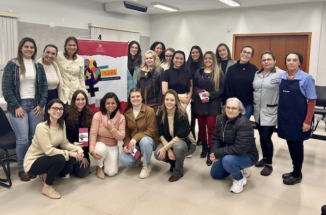 A imagem mostra um grupo de quinze mulheres posando para a foto em uma sala. Elas estão em pé e agachadas, e parecem estar em um ambiente de conferência ou reunião. No fundo, há um banner com a palavra "INDIRA" e o símbolo do feminino estilizado com cores vibrantes. As mulheres estão sorrindo e parecem participar de um evento ou encontro. Algumas seguram folhetos ou cadernos. A sala tem paredes claras e está iluminada.