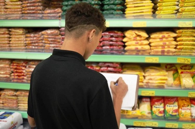 A imagem mostra um jovem aprendiz de  costas, usando uma camiseta preta, em um corredor de supermercado. Ele está segurando um caderno e uma caneta, fazendo anotações. Na frente dele, há prateleiras com pacotes de alimentos, aparentemente grãos e farináceos, como arroz ou feijão, organizados em fileiras. Os pacotes são de várias cores, predominantemente amarelos e vermelhos. Os preços estão visíveis nas prateleiras.
