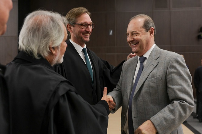 A imagem mostra três homens em um ambiente formal. Dois deles estão vestindo togas pretas e um está usando um terno cinza. Eles estão sorrindo e apertando as mãos, sugerindo um momento de cumprimento ou celebração. A cena ocorre em um ambiente interno, possivelmente em um local relacionado a eventos legais ou judiciais.