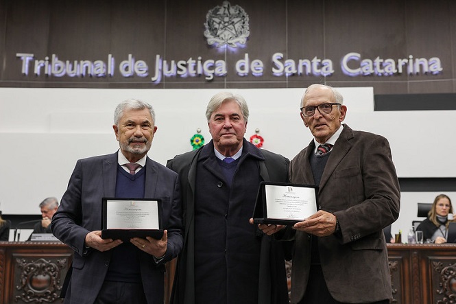 A imagem mostra três homens em pé em frente a um painel que diz "Tribunal de Justiça de Santa Catarina". Dois deles estão segurando placas de reconhecimento. Os homens estão vestidos formalmente com ternos e gravatas. Ao fundo, há um ambiente que parece ser uma sala de tribunal, com outras pessoas sentadas. A cena sugere um evento de premiação ou reconhecimento formal.
