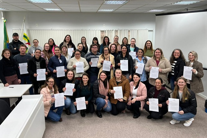 A imagem mostra um grupo de pessoas posando em uma sala de reunião. Muitas delas estão segurando folhas de papel. O fundo da sala tem bandeiras, e as pessoas parecem felizes e orgulhosas. Eles estão em duas fileiras, com algumas pessoas agachadas na frente para melhor visibilidade.