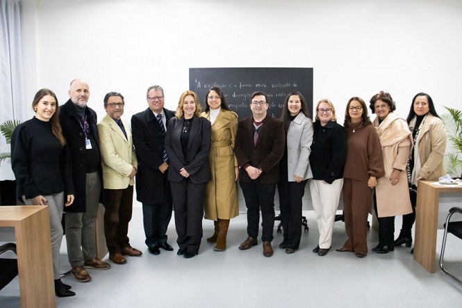 A imagem mostra um grupo de 13 pessoas posando para uma foto em uma sala. Eles estão organizados em uma única fileira. Ao fundo, há um quadro negro com uma citação escrita. A sala parece ser um ambiente formal, possivelmente em uma instituição educacional ou empresarial. As pessoas estão vestidas de maneira formal e há mesas nos cantos da sala.