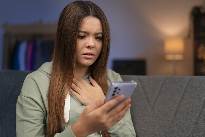 A imagem mostra uma mulher com expressão preocupada ou surpresa olhando para um celular. Ela está sentada em um sofá e usa uma blusa com capuz verde claro. Sua mão esquerda está no peito e ela parece estar em casa, devido à presença de uma lâmpada acesa ao fundo e um armário. A cena sugere que a mulher viu ou leu algo no telefone que a deixou intrigada e preocupada.