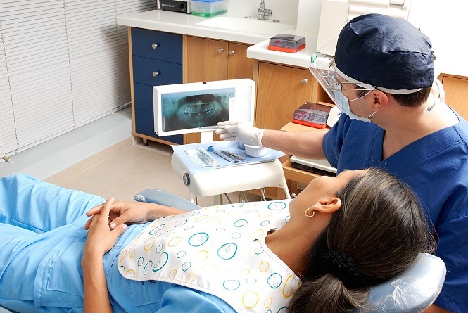 A imagem mostra um dentista sentado ao lado de um paciente em um consultório odontológico. O dentista está usando uniforme azul, máscara e luvas, e está mostrando uma radiografia dental ao paciente na tela de um computador. O paciente está deitado na cadeira, vestindo um avental de proteção. O ambiente parece ser limpo e organizado, com móveis em tons de madeira e equipamentos odontológicos.