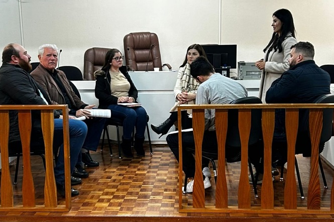 A imagem mostra quatro pessoas em um ambiente de escritório. Duas pessoas estão vestidas casualmente com blazers, e as outras duas estão em trajes formais, com ternos. Elas estão em pé lado a lado em uma sala que tem móveis de cor clara, incluindo um sofá e mesas de centro. Há decoração na parede e plantas ao redor, criando um ambiente profissional e acolhedor. Sobre a mesa ao centro, há livros e itens decorativos.