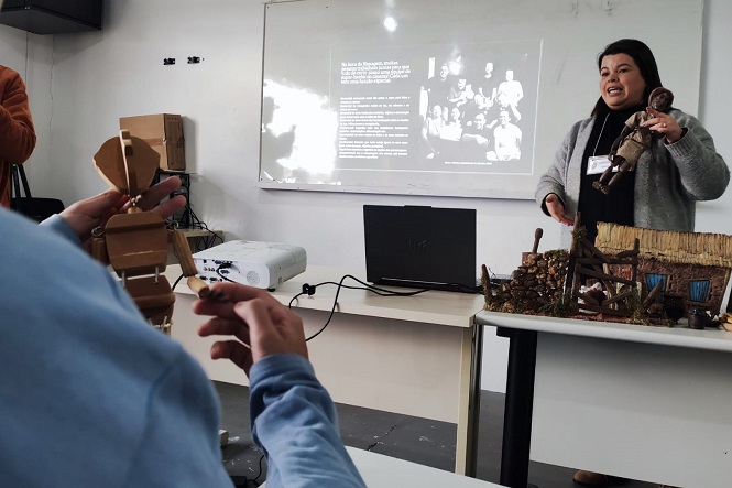 A imagem mostra uma sala de aula onde uma mulher está apresentando algo para um grupo. Ela segura uma boneca em uma mão e aponta para maquetes ou modelos de casas e paisagens que estão sobre a mesa. Ao fundo, existe uma tela de projeção com uma imagem em preto e branco de um grupo de pessoas e um texto, o que sugere que ela está fazendo uma apresentação ou explicando um conceito. Na frente, uma pessoa observa enquanto segura outro objeto nas mãos. Na mesa, há um projetor e um notebook. A cena parece ser uma aula ou oficina interativa.