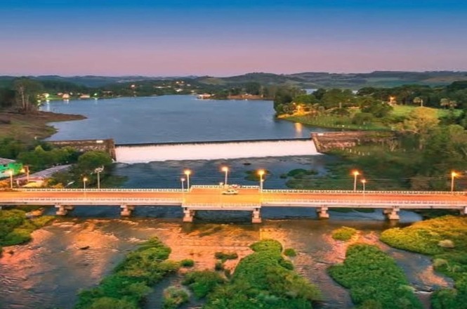 A imagem mostra uma paisagem com uma ponte iluminada atravessando um rio com vegetação ao redor. No fundo, há uma barragem que retém uma grande quantidade de água formando um lago. O cenário é cercado por árvores e colinas distantes, sob um céu azul-rosado típico do entardecer. As luzes da ponte refletem na água, criando uma atmosfera tranquila e pitoresca.