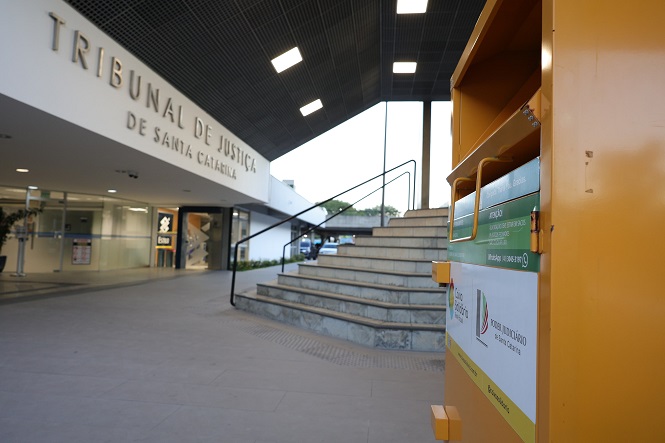 A imagem mostra a entrada do "Tribunal de Justiça de Santa Catarina". Em primeiro plano, à direita, há um contêiner ou caixa de coleta de cor amarela, que possui adesivos e informações. Ao fundo, é possível ver escadas que levam à entrada principal do prédio, que é moderno e possui um teto com iluminação embutida. A fachada é clara e apresenta o nome do tribunal em letras grandes.
