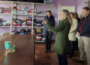 A imagem mostra um grupo de pessoas em uma sala. Na parede do fundo, há prateleiras cheias de roupas dobradas. À frente, uma mesa de madeira com um brinquedo azul em cima. As pessoas estão em pé, provavelmente ouvindo alguém falar ou apresentando algo. Elas parecem estar em um ambiente acolhedor ou em uma instituição que organiza doações de roupas.