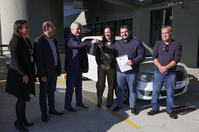 A imagem mostra seis pessoas em uma cerimônia de entrega de um carro. Todas estão de pé em frente ao veículo branco, que está estacionado em uma área externa. Duas das pessoas no centro estão se cumprimentando, e uma delas está segurando um conjunto de chaves. Outra pessoa segura um documento, possivelmente relacionado à entrega do carro. O grupo parece estar em um ambiente profissional ou institucional, dada a vestimenta formal de alguns indivíduos. Há um clima de celebração ou formalidade no ar.
