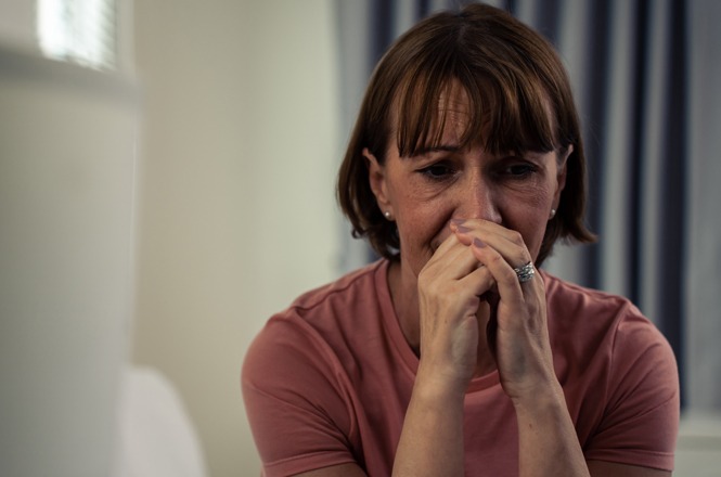 A imagem mostra uma mulher aparentando estar emocionalmente angustiada e preocupada. Ela está vestindo uma camisa rosa, com as mãos perto do rosto e uma expressão que sugere tristeza ou preocupação. O fundo parece ser um ambiente interno, com cortinas azuis visíveis.