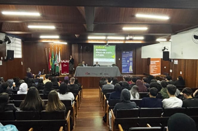 A imagem mostra um auditório com várias pessoas sentadas assistindo a uma apresentação. À frente, há uma mesa com dois palestrantes, e atrás deles há um telão com o título "Oficina: Escolher Peças de Santos Cristais". Na lateral, há banners informativos e bandeiras. O ambiente é fechado, com um teto de madeira e iluminação com luzes fluorescentes.