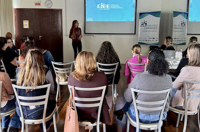 A imagem mostra uma sala onde várias pessoas estão sentadas em cadeiras, assistindo a uma apresentação. Na frente, uma mulher está de pé, falando para o público. Há um projetor exibindo o título "Centro de Ensino e Educação" e banners laterais com a inscrição "Novos Caminhos". A sala tem piso de madeira e as cadeiras estão organizadas em fileiras. As pessoas estão prestando atenção à apresentação.