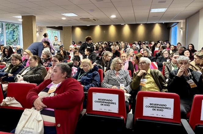 A imagem mostra um auditório cheio de pessoas sentadas, a maioria das quais parece estar atenta a um evento ou apresentação. Algumas cadeiras nas fileiras da frente têm placas indicando "RESERVADO COORDENADORES DE EIXO". As pessoas parecem estar em um ambiente interno, com iluminação de teto e grandes janelas ao fundo. A atmosfera sugere um encontro, como uma conferência ou palestra. Muitas pessoas estão usando crachás, o que indica que é um evento oficial. A sala parece bem iluminada e organizada.