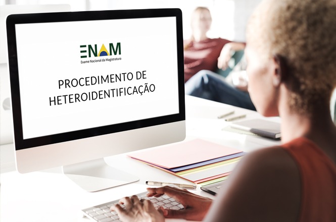 A imagem mostra uma pessoa sentada em uma mesa usando um computador. Na tela do computador, está exibido o texto "ENAM Exame Nacional da Magistratura" e "Procedimento de Heteroidentificação". Em segundo plano, há outra pessoa sentada em um sofá. Há itens como cadernos ou papéis em cima da mesa ao lado do computador.