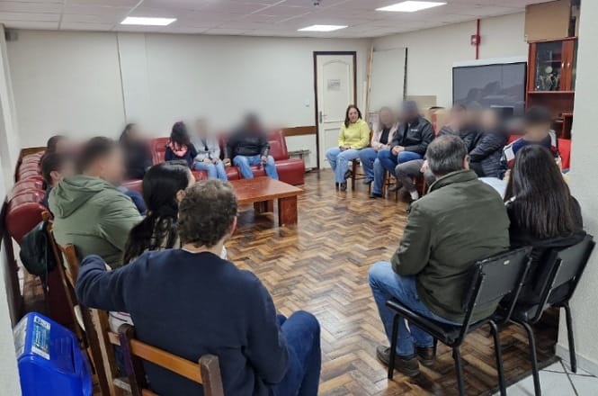 A imagem mostra um grupo de pessoas sentadas em um círculo em uma sala. As pessoas estão em cadeiras e sofás, e parecem estar participando de uma reunião ou discussão em grupo. O piso é de madeira, há uma mesa de centro no meio, e a sala tem iluminação de teto. Ao fundo, há uma porta e uma mobília, possivelmente uma estante ou armário.