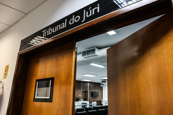 A imagem mostra uma entrada de madeira com portas duplas abertas que levam a uma sala de tribunal. Acima das portas, há uma placa preta com letras brancas que diz "Tribunal do Júri". No interior, é possível ver parte de uma sala com mesas, cadeiras e equipamentos eletrônicos, tipicamente associados a um ambiente de julgamento.
