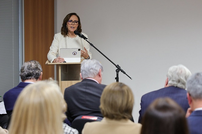 Na imagem, há uma mulher vestida de traje formal falando ao microfone, em um púlpito. Ela segura um tablet enquanto faz uma apresentação ou discurso. À sua frente, há várias pessoas sentadas, todas de costas, aparentemente ouvindo atentamente. O ambiente parece ser uma sala de conferências ou reunião.
